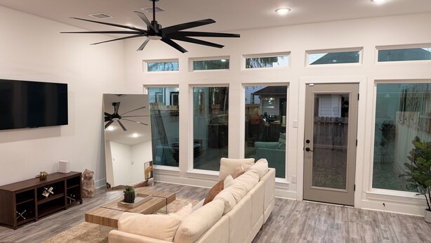 A modern living room with a large sofa, a coffee table, and a TV. There are multiple windows and a sliding glass door.