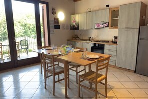 A dining area with a table set for four, a kitchen with light wood cabinets, and a view through a glass door to an outdoor area. - Free Park and Swimming Pool, Centre of Peschiera (Peschiera del Garda)