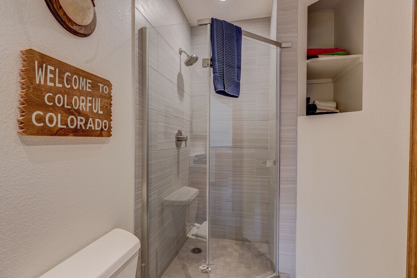 Un baño moderno con cabina de ducha de vidrio, inodoro blanco y barra para toallas. Se ve un cartel de bienvenida a Colorado en la pared.