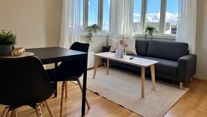 A modern living room with a dining table, black chairs, a grey sofa, and a coffee table. There are two windows with white curtains, a small plant on the windowsill, and a rug on the floor. - Forenom Serviced Apartments Stavanger (Stavanger)