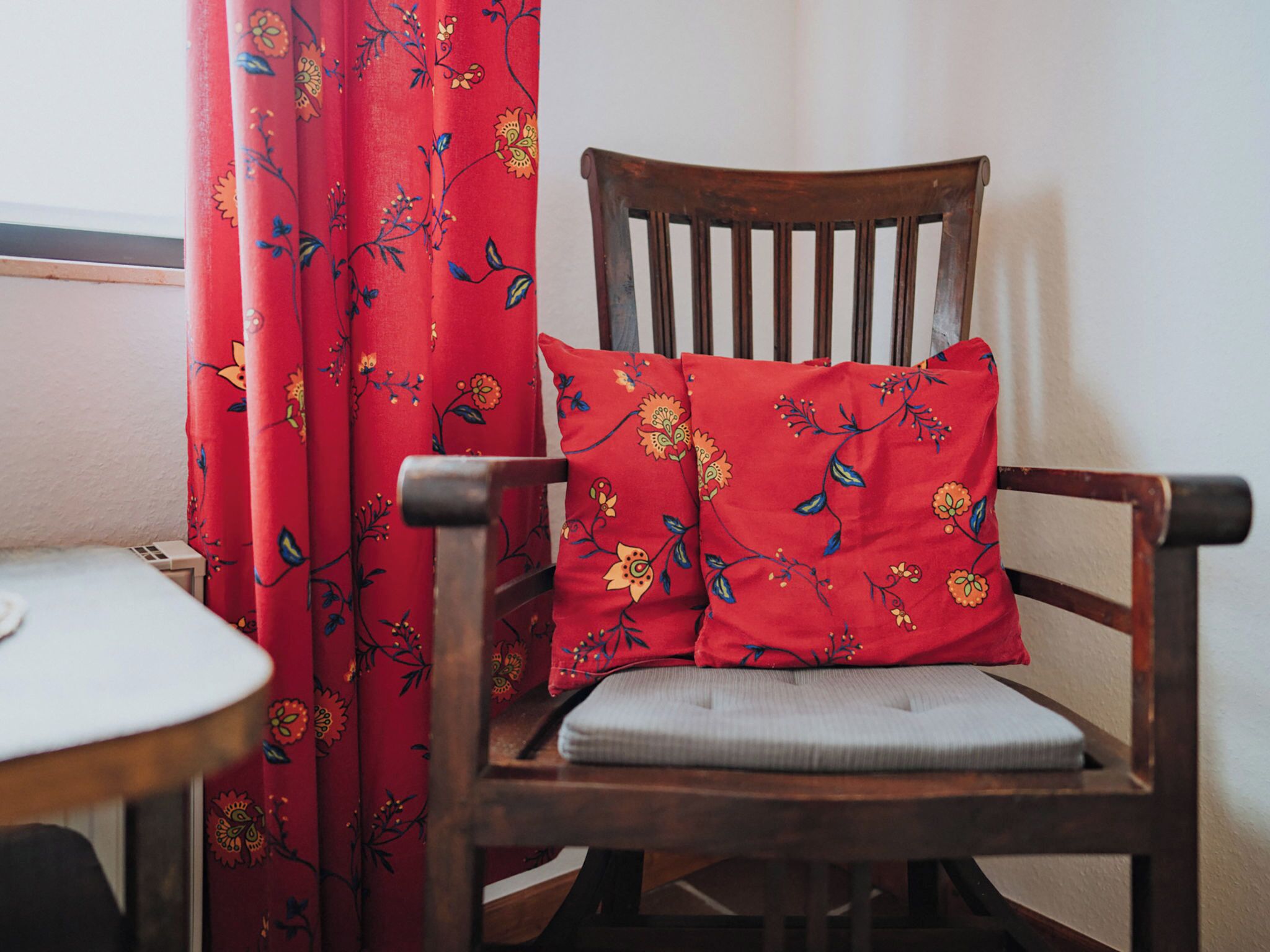 A wooden chair with a cushion and a floral-patterned pillow, positioned next to a table and a red curtain with a floral design.