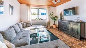A modern living room with a wooden ceiling, a grey sofa, a glass coffee table, a TV mounted on the wall, and a kitchen area with a dark cabinet. - Teutoburger Wald 1 by Interhome (Schwalenberg)