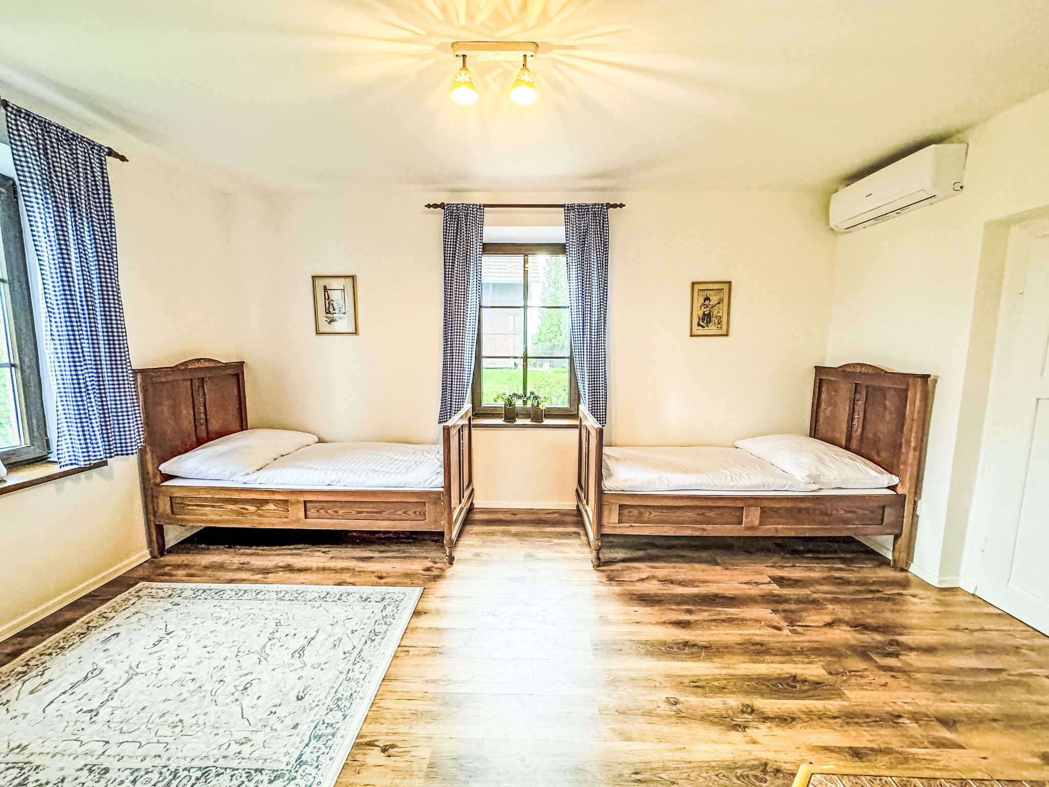 Une chambre avec deux lits en bois, une fenêtre avec des rideaux, une petite table avec une plante et un appareil de climatisation.