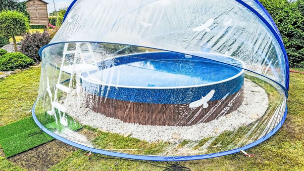 A transparent dome enclosure with a blue interior, surrounded by a grassy area and a wooden structure in the background. - Rabštejn by Interhome (Zihle)