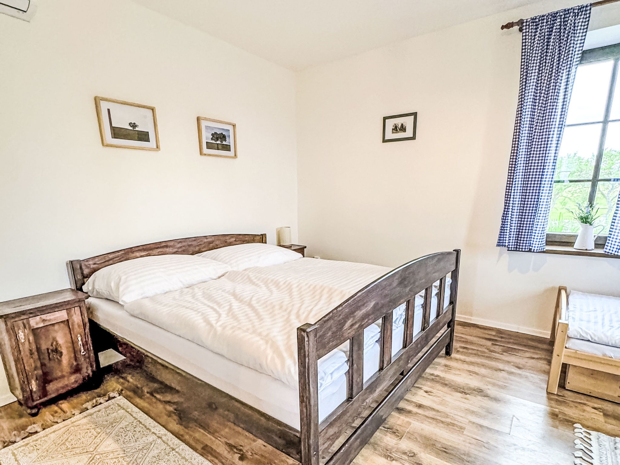 A bedroom with a wooden bed, a nightstand, a window with checkered curtains, and three framed pictures on the wall.