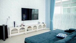 Ein modernes Schlafzimmer mit einem großen Flachbildfernseher an der Wand, einem weißen Konsolentisch mit Schubladen, einem blauen Sofa und einem großen Fenster mit Vorhängen.