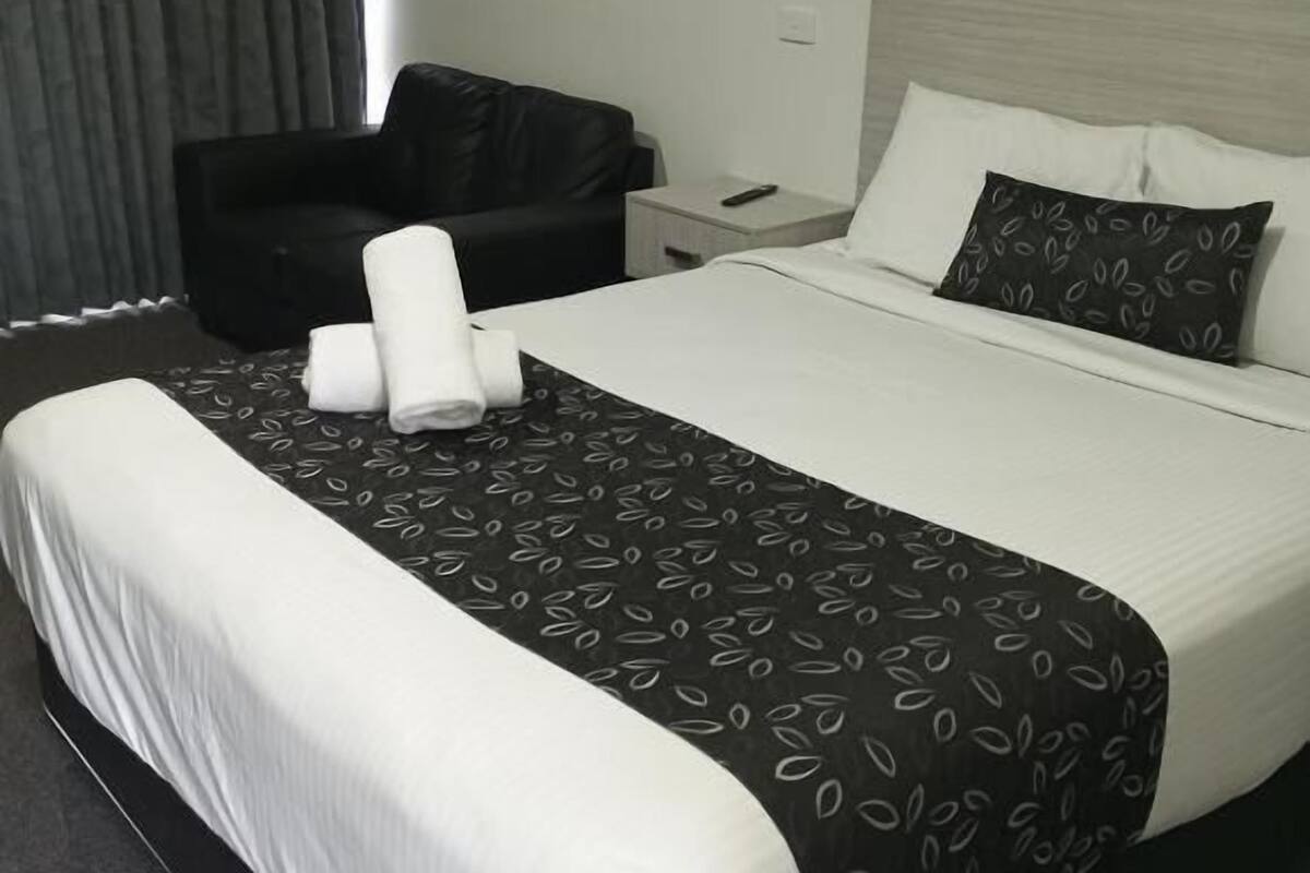Una habitación de hotel con una cama grande, un sillón de cuero negro, una mesita de noche con un control remoto y una ventana con cortinas.