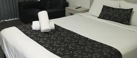A hotel room with a large bed, a black leather chair, a bedside table with a remote control, and a window with curtains.
