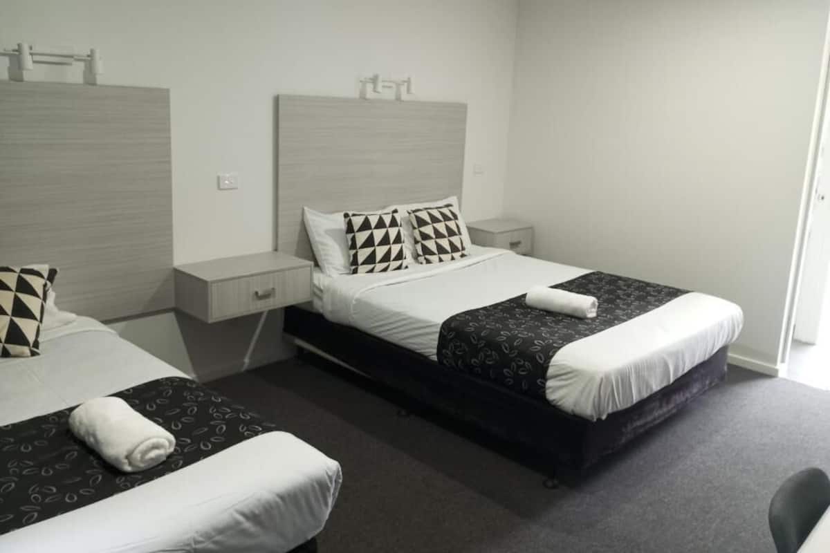 Habitación de hotel con dos camas, cada una con almohada y manta. Se observa un mesita de noche con lámpara y cajón.