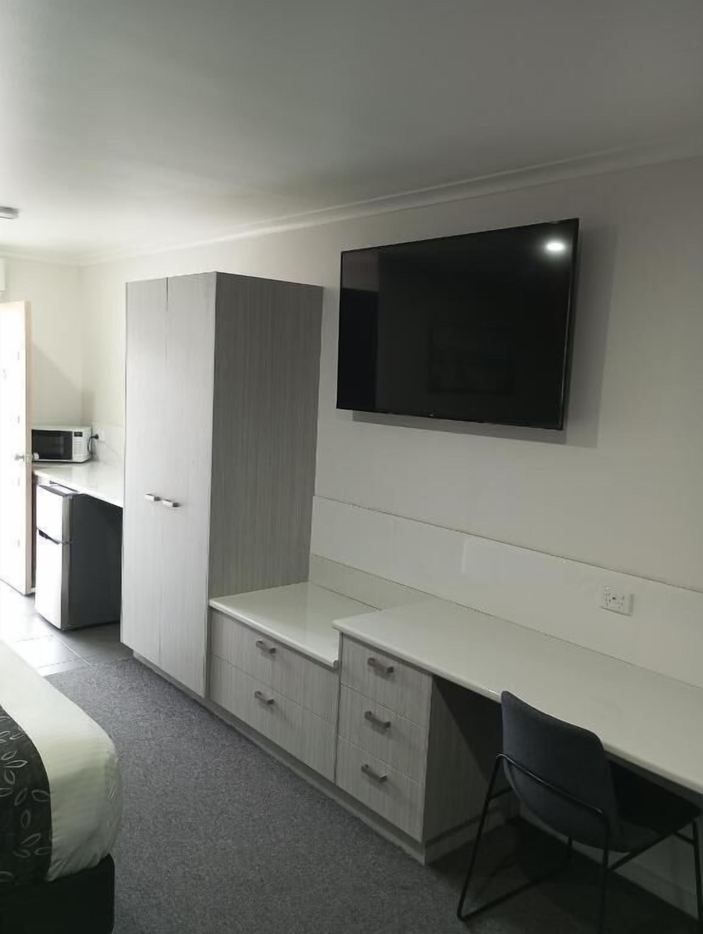 A modern, compact room with a flat-screen TV mounted on the wall, a kitchenette area, and a desk with a chair.