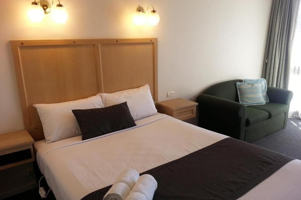 Habitación de hotel con una cama grande, dos mesitas de noche, un sillón verde y luces fijadas a la pared.