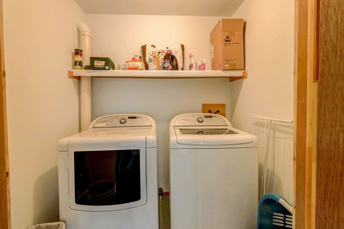 A laundry room with two front-loading washing machines and a shelf above containing cleaning supplies.