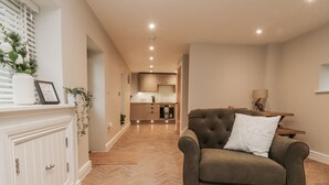 A living room with a sofa, a fireplace, a kitchen in the background, and a herringbone floor pattern. - Henside Cottage (Settle)