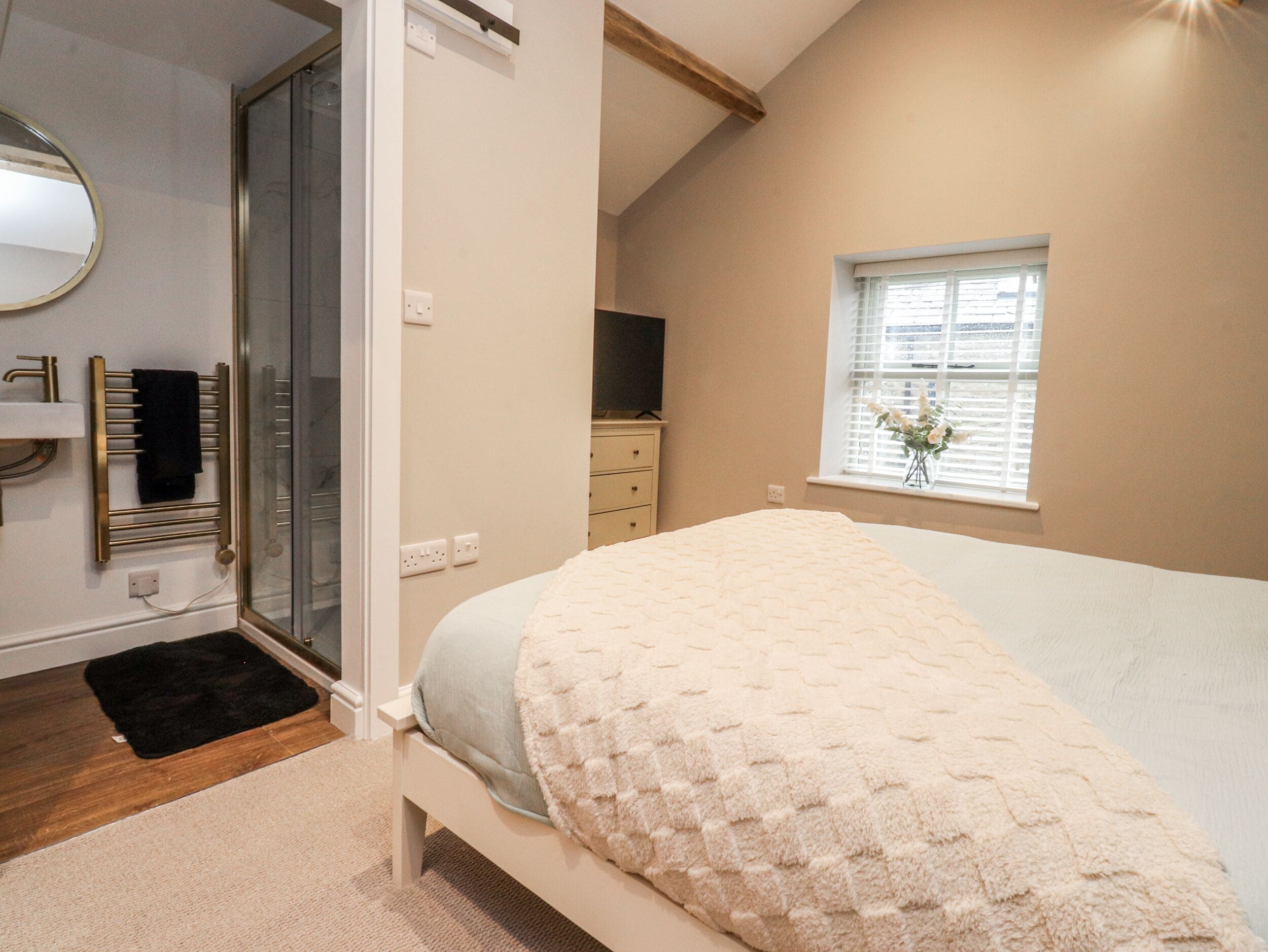 A bedroom with a bed, a bedside table, a mirror, a window with blinds, and a door leading to a bathroom.
