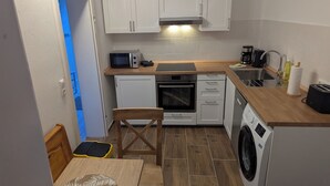 Una cocina compacta con armarios blancos, microondas, horno, lavadora y una mesa de madera con una silla.