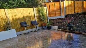 A patio area with a tiled floor, two black bar stools, a wooden fence, and potted plants. - 3 bed House, Sleeps 5, Near Airport and Town, Outside Parking Free (Manchester)