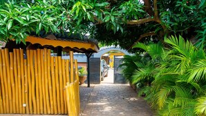 A paved pathway leading to a building with a bamboo fence and tropical plants. - Hôtel Ô Balkan (Lomé)