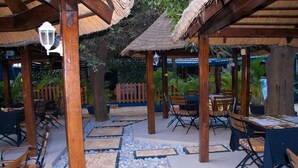 An outdoor dining area with wooden tables and chairs, thatched roof structures, and a tiled floor with a pebble pattern. - Hôtel Ô Balkan (Lomé)