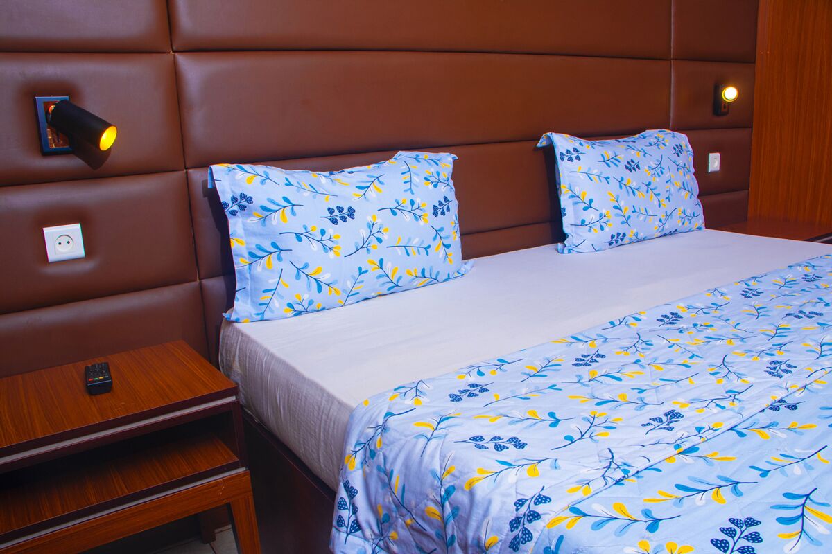 Habitación de hotel con una cama grande, dos almohadas de estampado floral, una mesita de noche con un control remoto y una lámpara de pared.