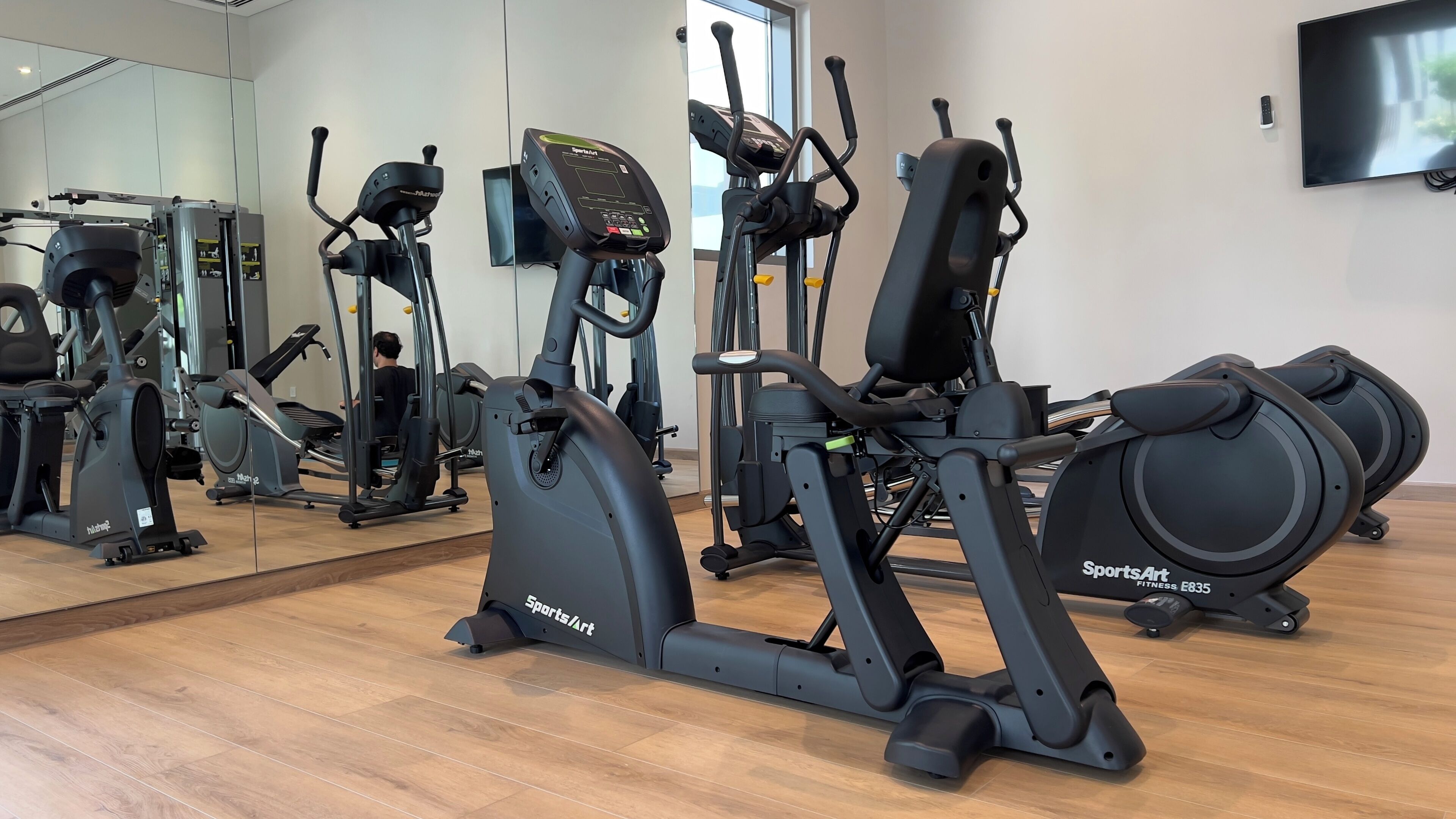Une salle de sport équipée de nombreux appareils d’exercice, notamment des elliptiques et un vélo d’appartement, disposés dans un espace vaste agrémenté d’un grand miroir et d’un téléviseur mural.