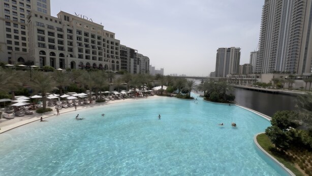 Una amplia piscina al aire libre rodeada de palmeras y sillas de descanso, con rascacielos al fondo.