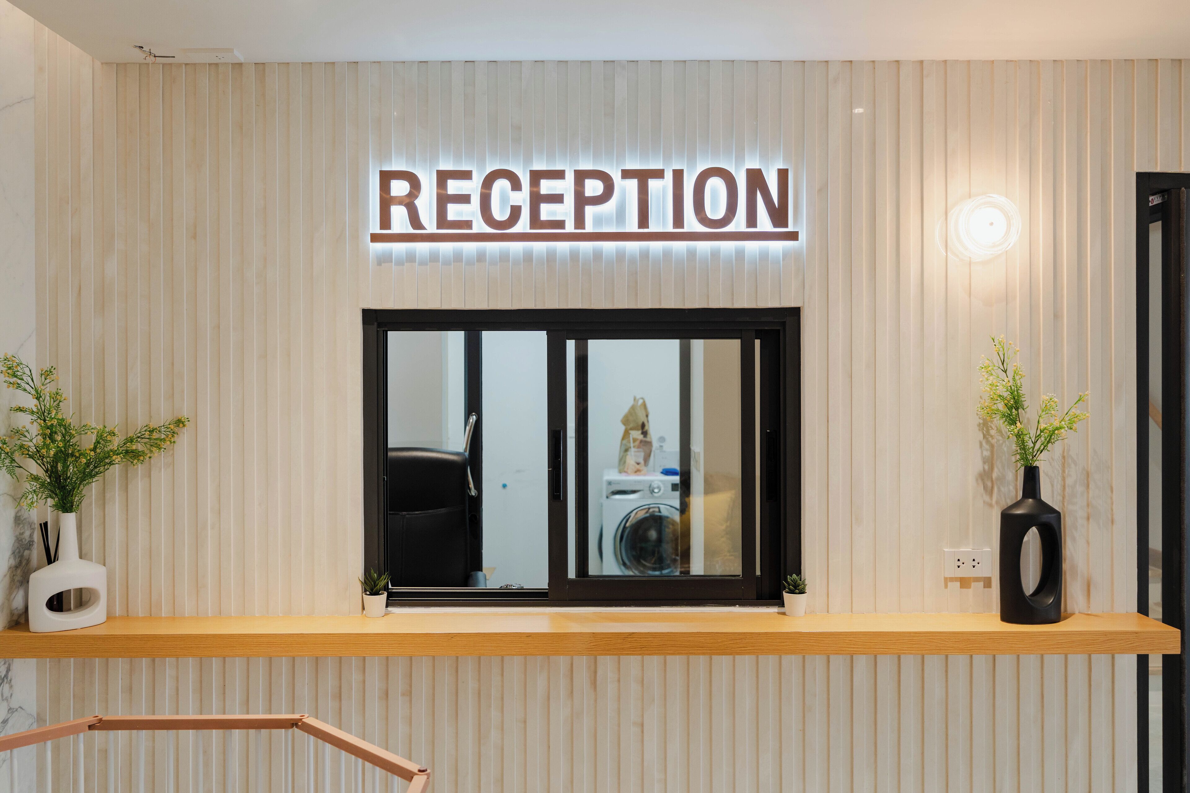 Ett receptionsområde med en skylt som visar ordet ”RECEPTION” ovanför ett fönster. På var sin sida om receptionsdisken står två krukväxter.