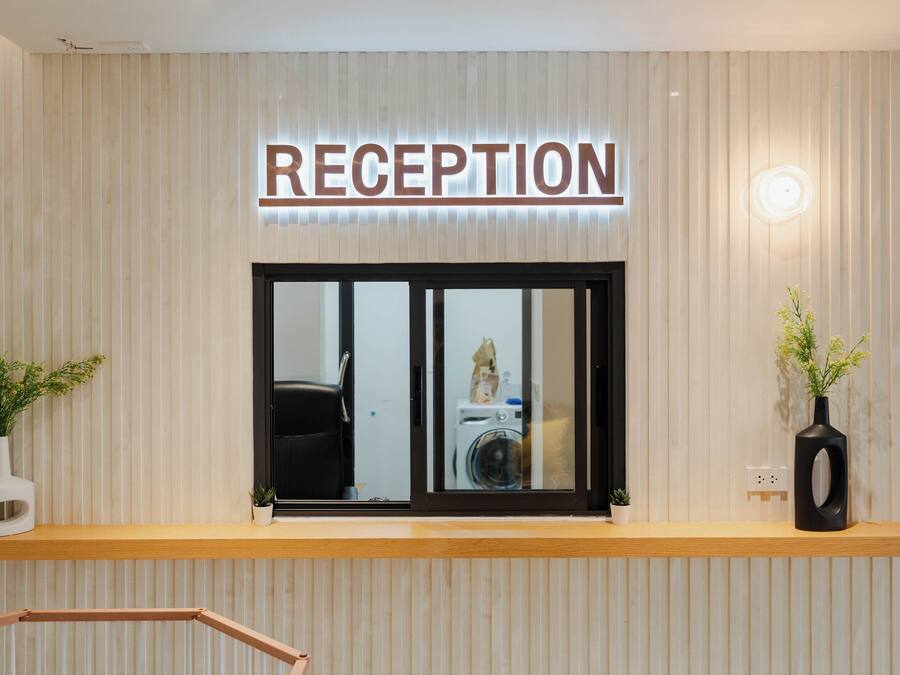 Ett receptionsområde med en skylt som visar ordet ”RECEPTION” ovanför ett fönster. På var sin sida om receptionsdisken står två krukväxter.