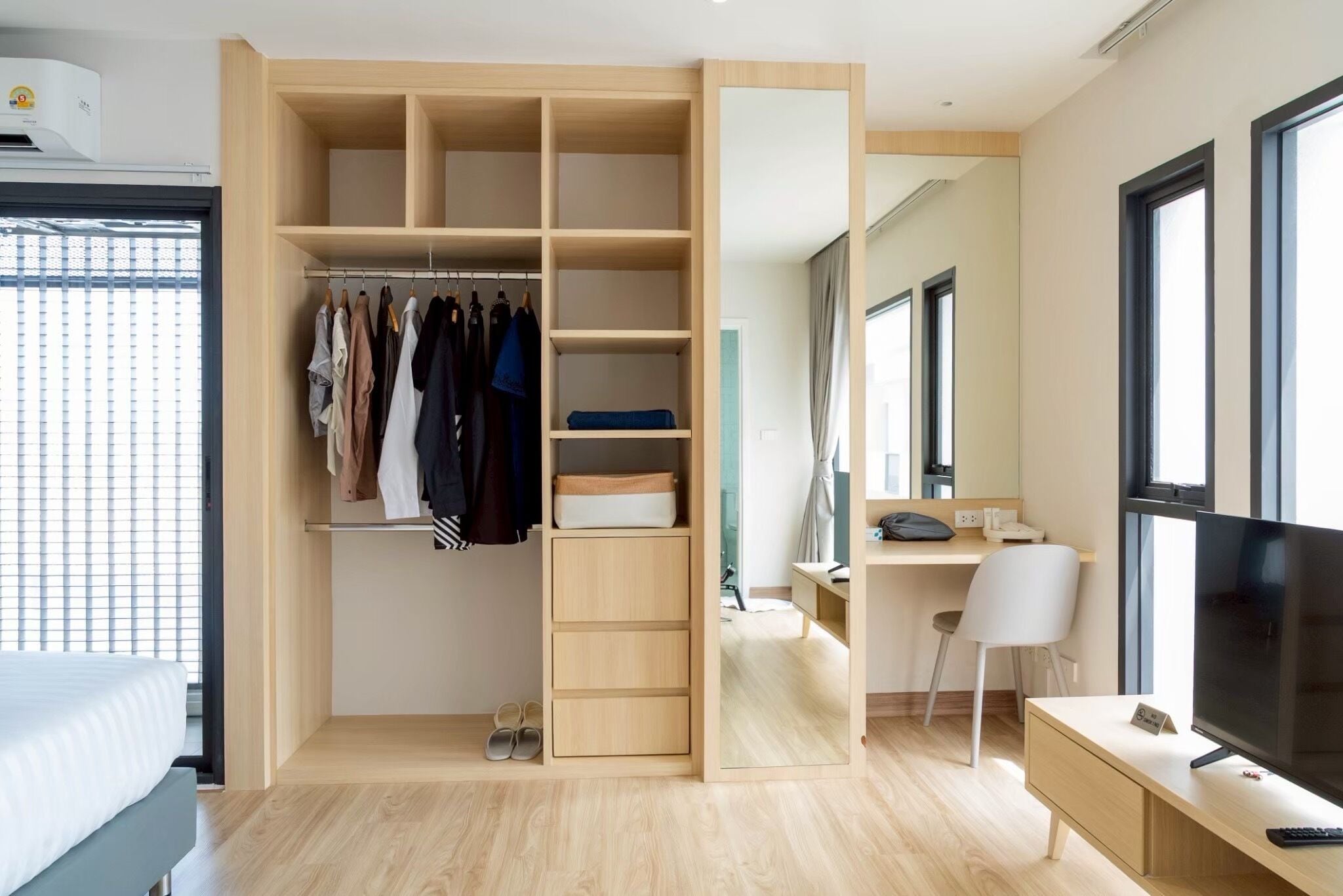 A modern hotel room with a walk-in closet, a desk with a chair, a television, and a large window.