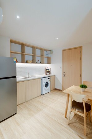 A compact kitchen with a refrigerator, washing machine, wooden cabinets, and a dining area with a table and chairs. - I-Zen Residence Central Eastville (Bangkok)