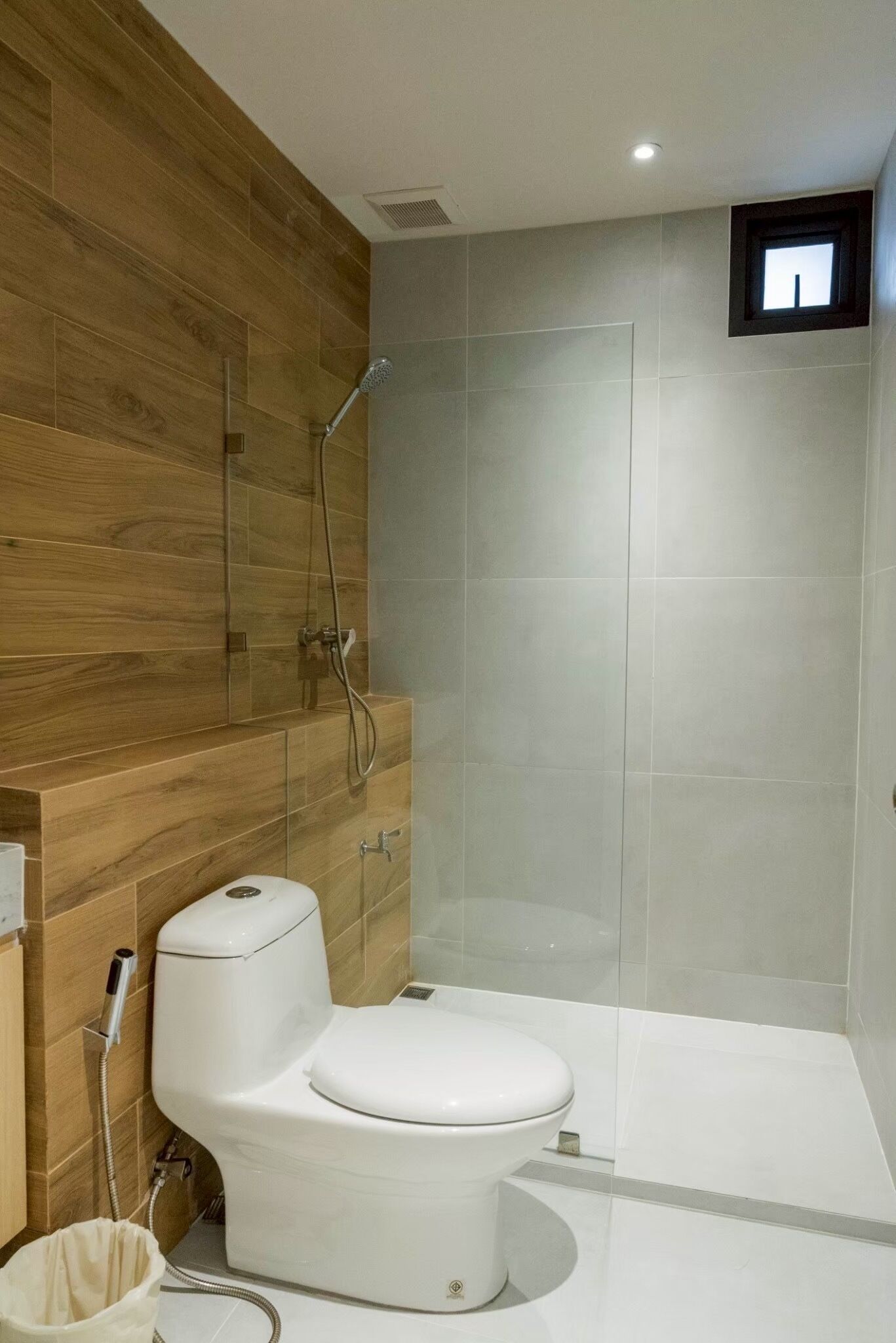 A modern bathroom with a wooden wall, a white toilet, a shower enclosure, and a small window.