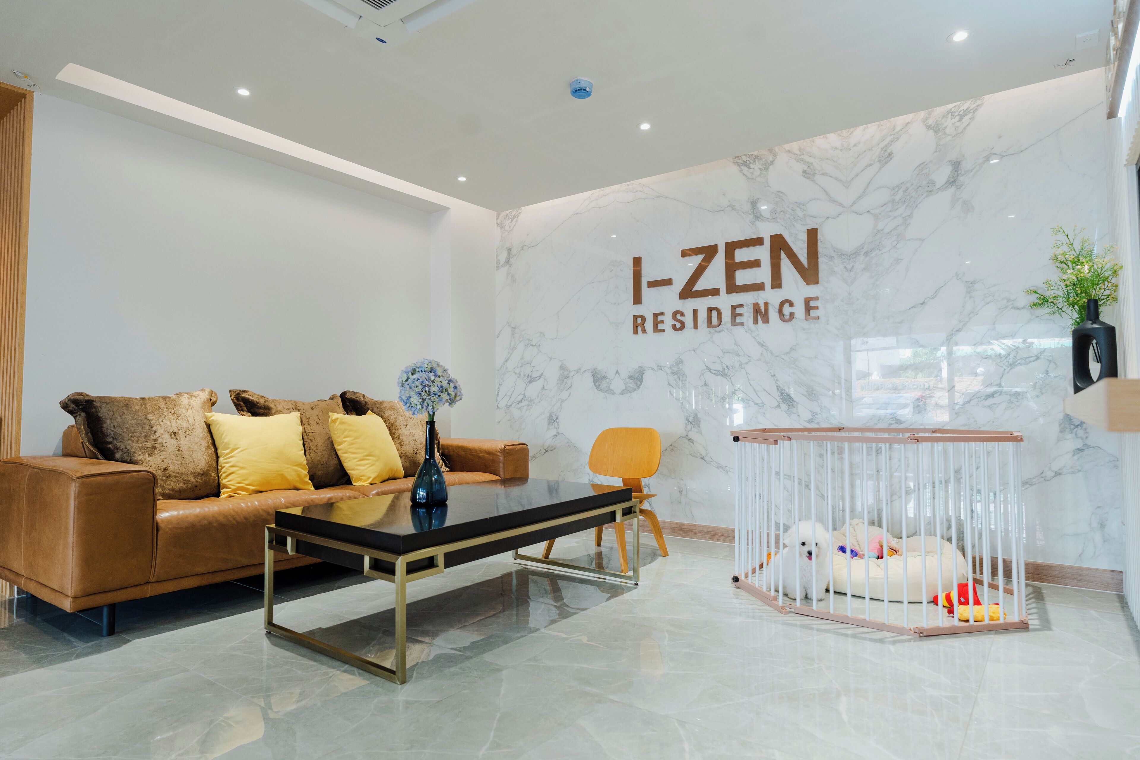 A modern living room with a sofa, coffee table, and a pet bed. The room has a marble wall with the text "I-ZEN RESIDENCE" and a ceiling with recessed lighting.