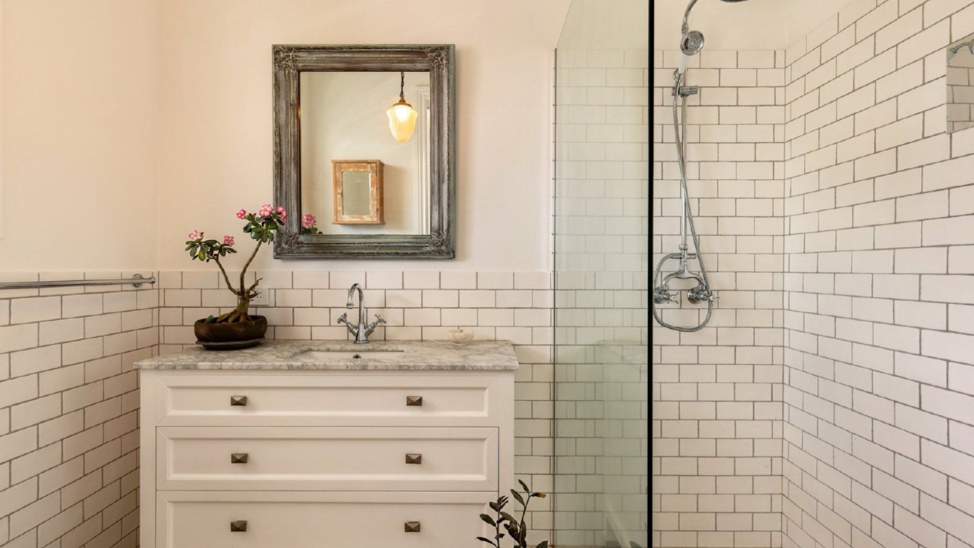 A bathroom with a white vanity, a mirror with a picture frame, a potted plant, and a shower with a glass door.