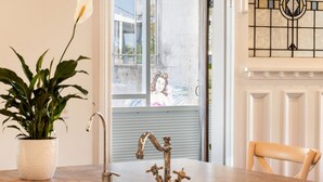 Una habitación con una planta en maceta, un grifo metálico, una silla y una ventana que da a un edificio.