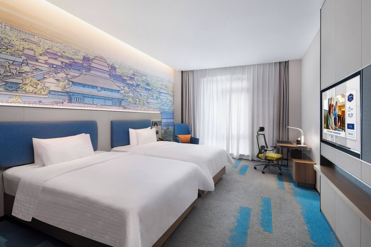 A hotel room with two beds, a desk, a chair, a large map on the wall, and a TV screen displaying room information.