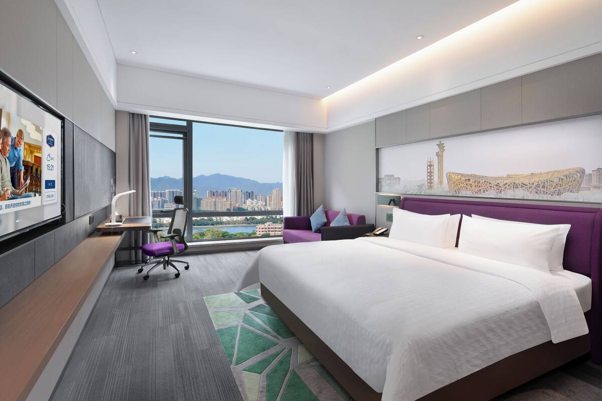 A hotel room with a large bed, a desk, a chair, a view of the city, and a window.