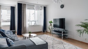 A modern living room with a grey sofa, a small round table, a TV on a stand, and a large window with curtains. - Spacious 2BR City Retreat with Sauna (Helsinki)