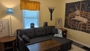 Una sala de estar con un sofá, una mesa de centro, una lámpara, un ventanal con persianas, un tapiz y una obra de arte colgada en la pared.