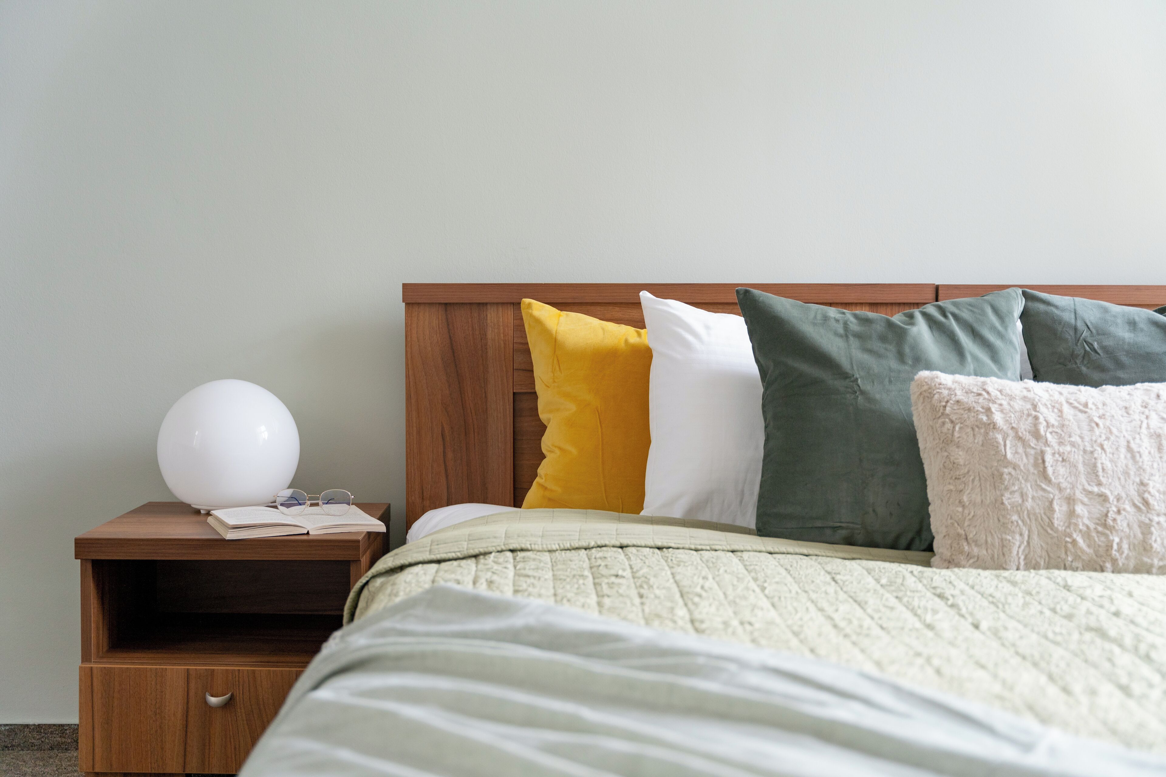 A neatly made bed with a quilted blanket, a wooden nightstand with a lamp, and a bedside table with a book and glasses.