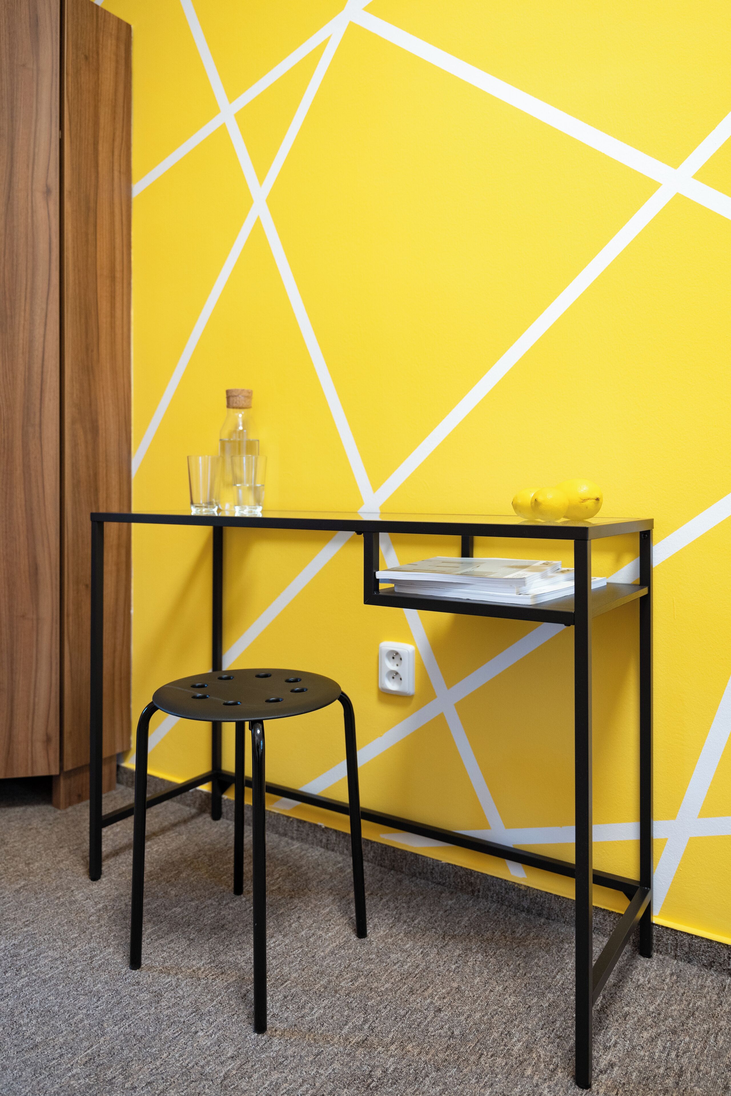 A small, black metal table with a stool, a cork bottle, and a glass on it, against a yellow wall with white geometric patterns.