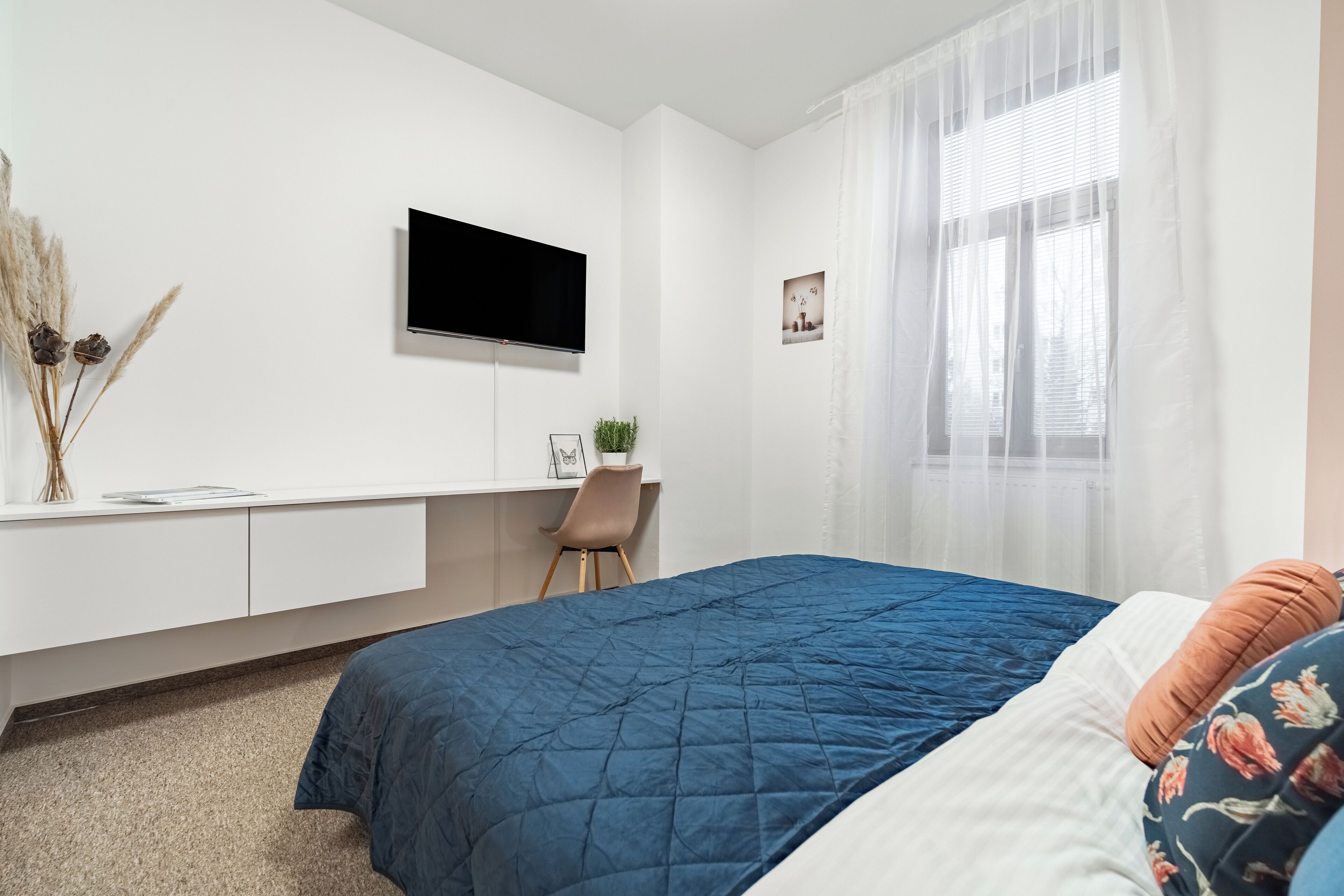 Une chambre à coucher avec un lit, un bureau, une télévision et une fenêtre ornée de rideaux légers.