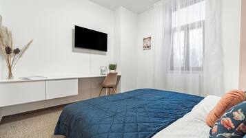 Une chambre à coucher avec un lit, un bureau, une télévision et une fenêtre ornée de rideaux légers.