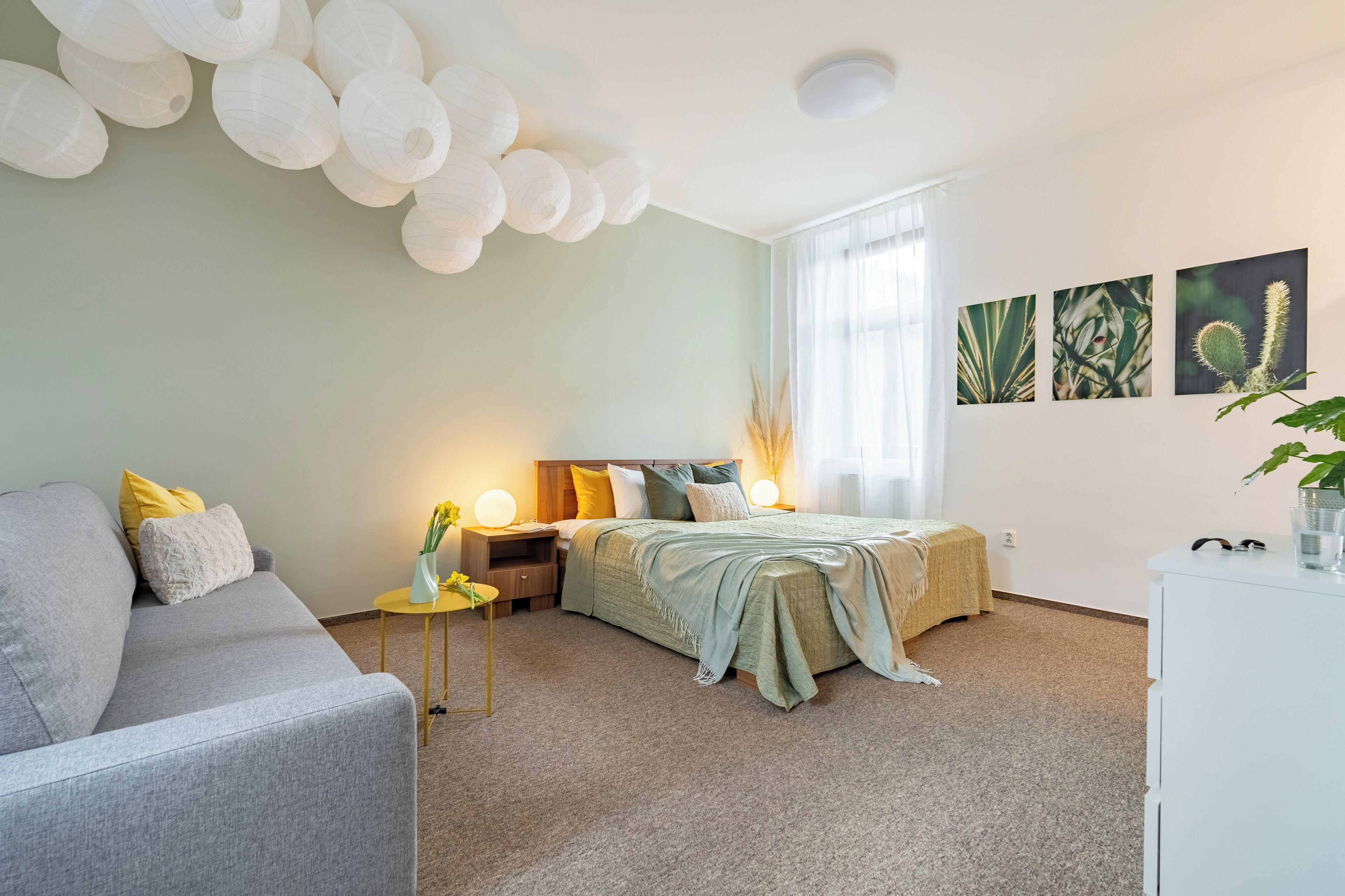 A modern bedroom with a grey sofa, a bed with a green blanket, bedside tables with lamps, and wall art depicting plants.