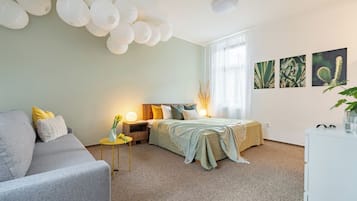 A modern bedroom with a grey sofa, a bed with a green blanket, bedside tables with lamps, and wall art depicting plants.