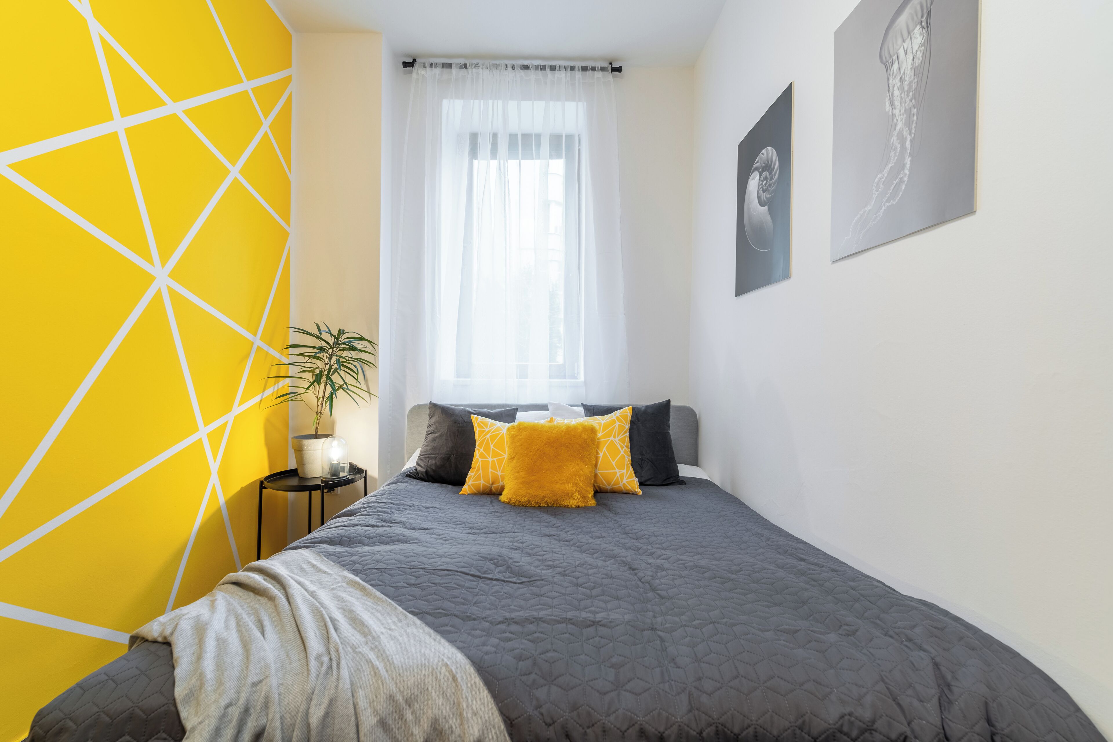 A bedroom with a bed, a window with sheer curtains, a potted plant, and artwork on the wall.