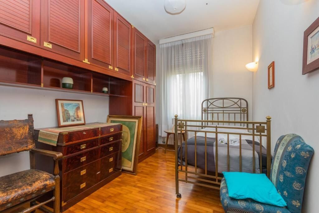 A room with a wooden cabinet, a bed with a metal frame, a blue patterned chair, and a wooden chair with a worn seat.