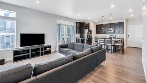 A modern living room with a grey sofa, a flat-screen TV, and a dining area with a kitchen in the background. - Comfort Stay w/ Studio • Thanksgiving Point (Lehi)