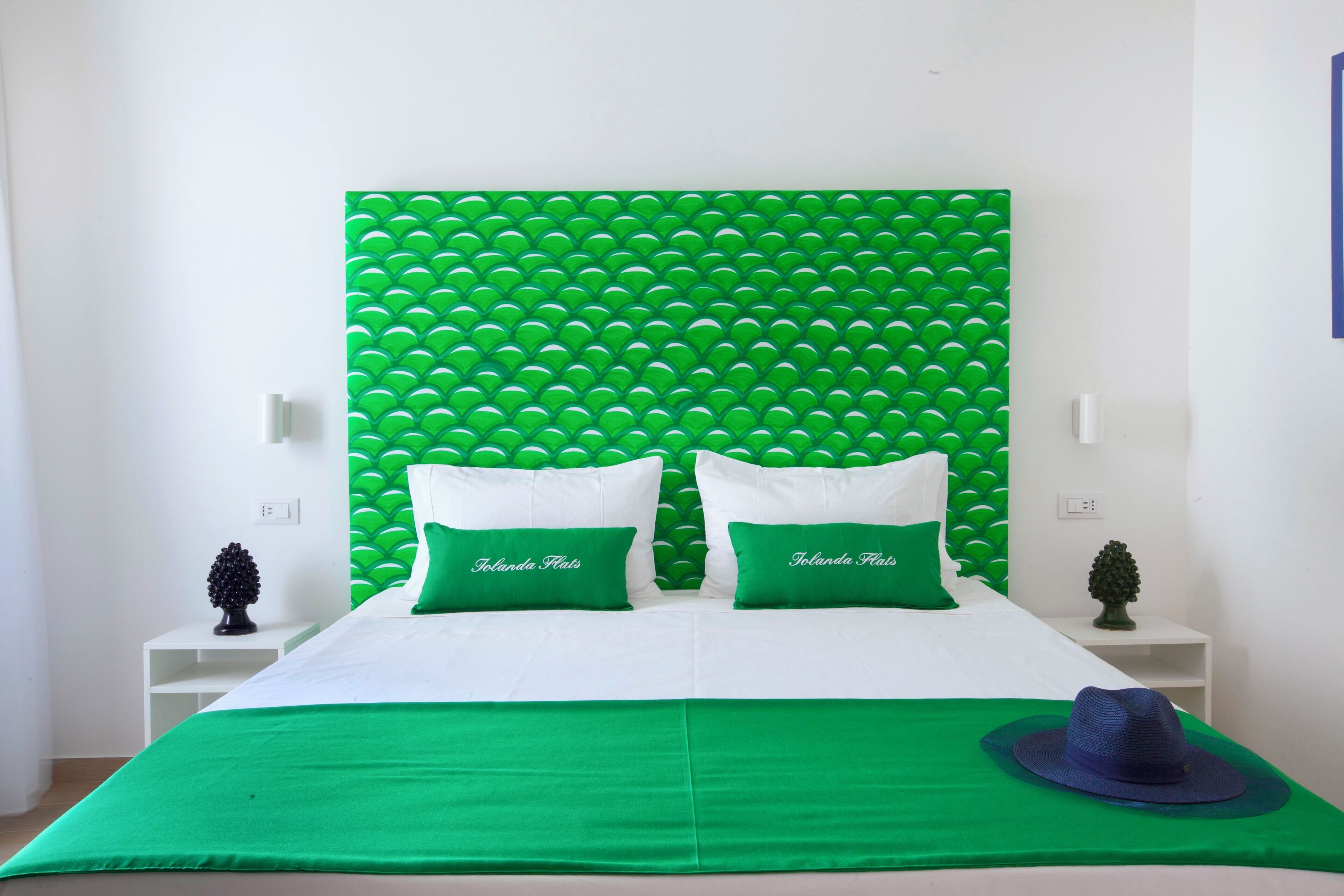 A bed with a green and white color scheme, two pillows with the text "Tolandu Flats," a green bedspread, and a blue hat on the bed.