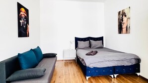 Ein modernes Schlafzimmer mit grauem Sofa, einem Bett mit grauer Bettdecke, Holzboden und zwei an der Wand befestigten Kunstwerken.