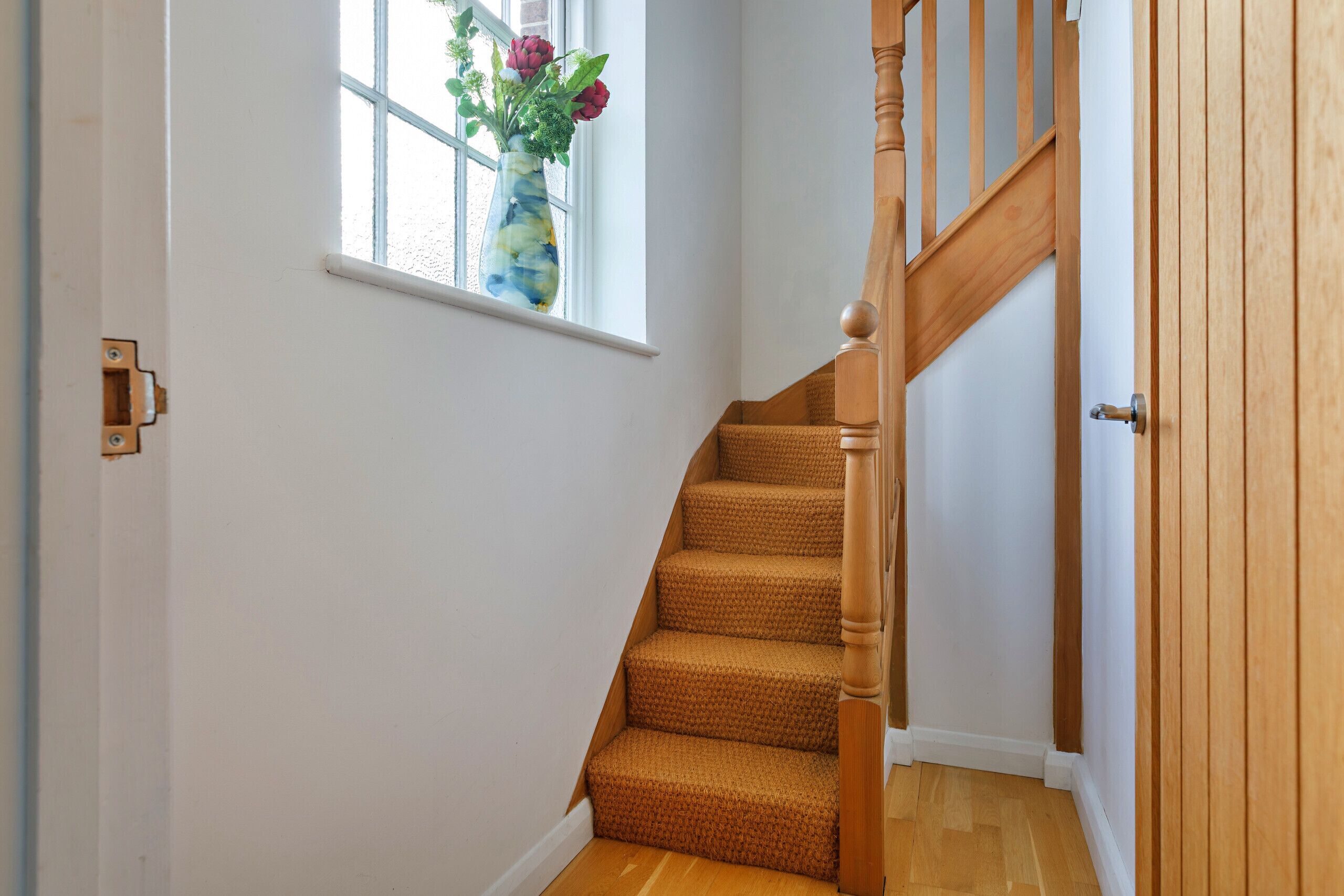 A wooden staircase with a handrail, leading to a door. A window with a vase of flowers is visible on the wall.