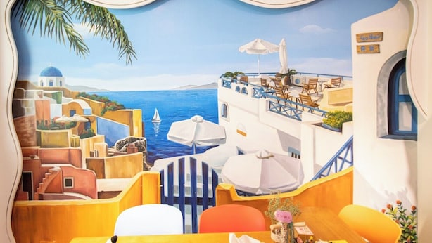 A mural of a coastal scene with a view of the sea, a sailboat, and a building with outdoor seating and umbrellas. - GOHOME (Kaohsiung)